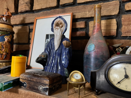 a stone fireplace mantle with a wizard statue, painted bottle, framed picture, clock, a globe timepiece, wooden box, ornamental stein, etc are visible